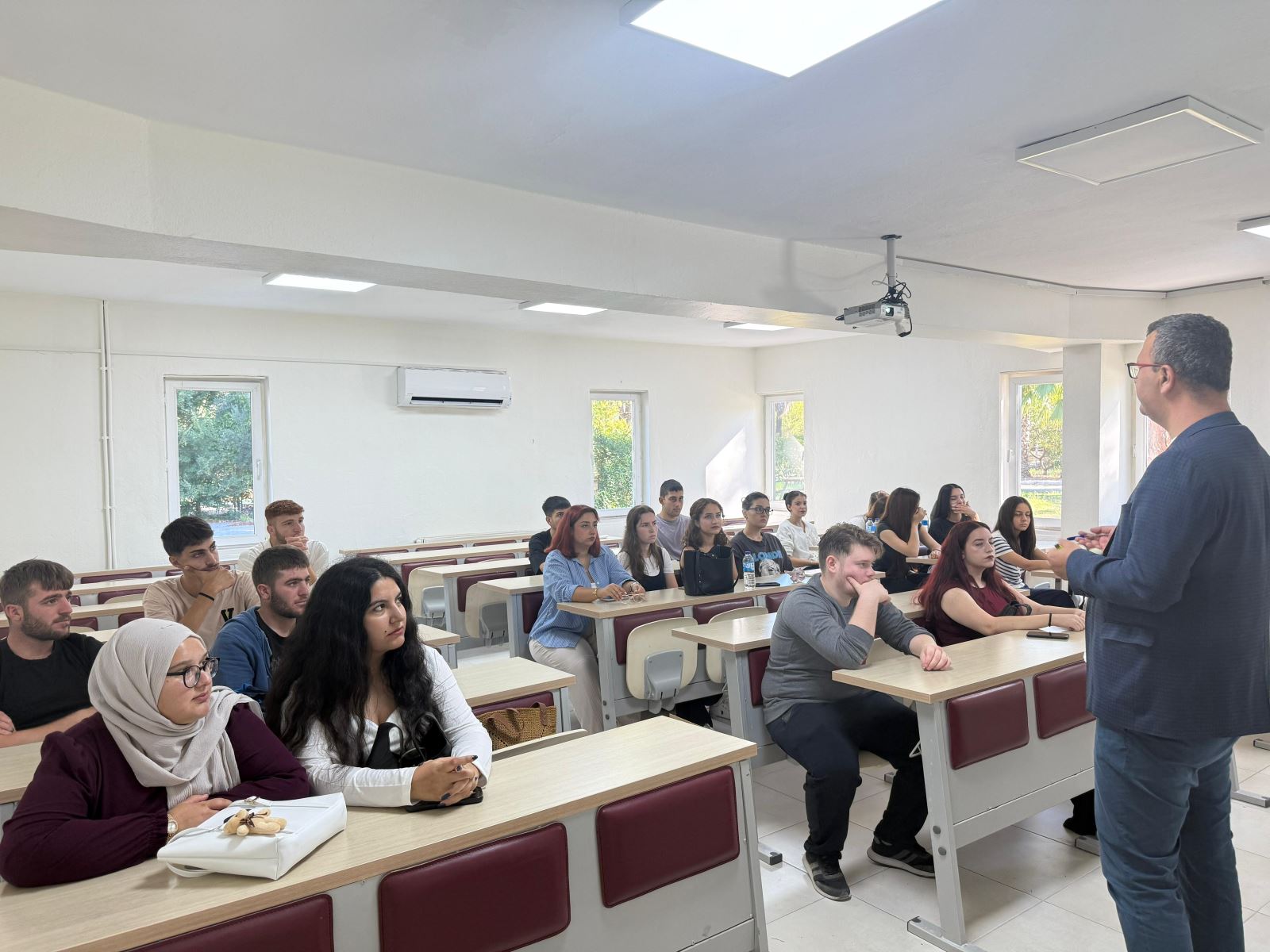 Derslikte bir öğretim elemanı öğrencilere hitap ediyor; sıralarda oturan öğrenciler dikkatle dinliyor, aydınlık sınıf ortamı.